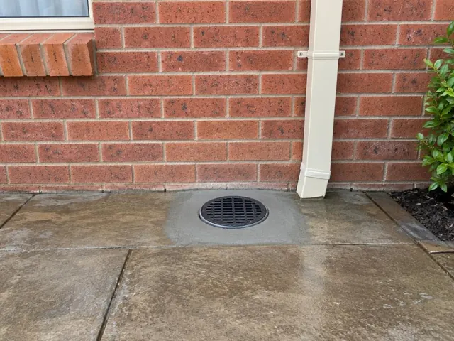 Clean outdoor drain grate in wet concrete paving beside a brick wall in Eagleton